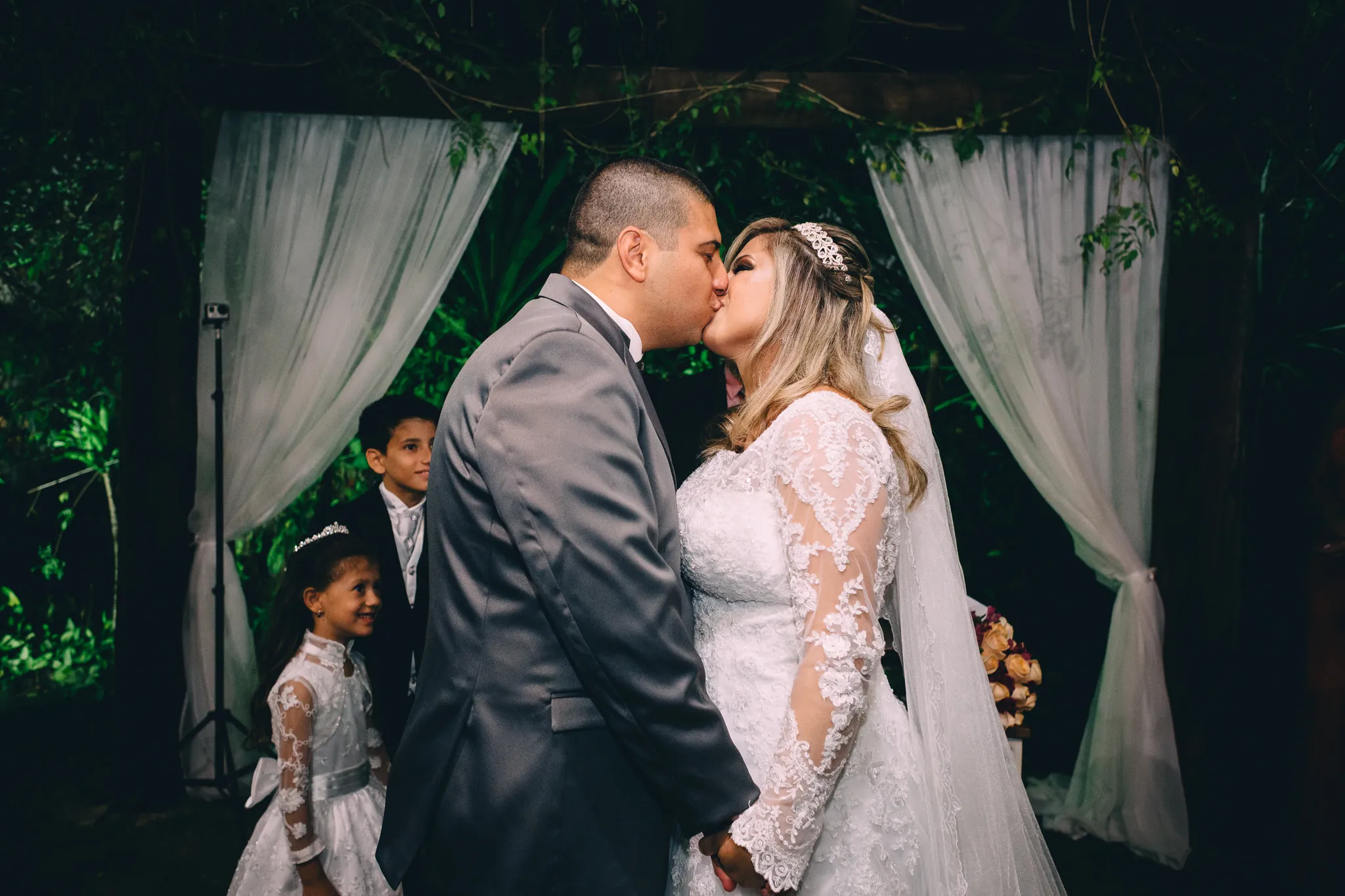 Gislaine e Thiago se beijando sob o arco de cortinas, com as crianças da família ao lado