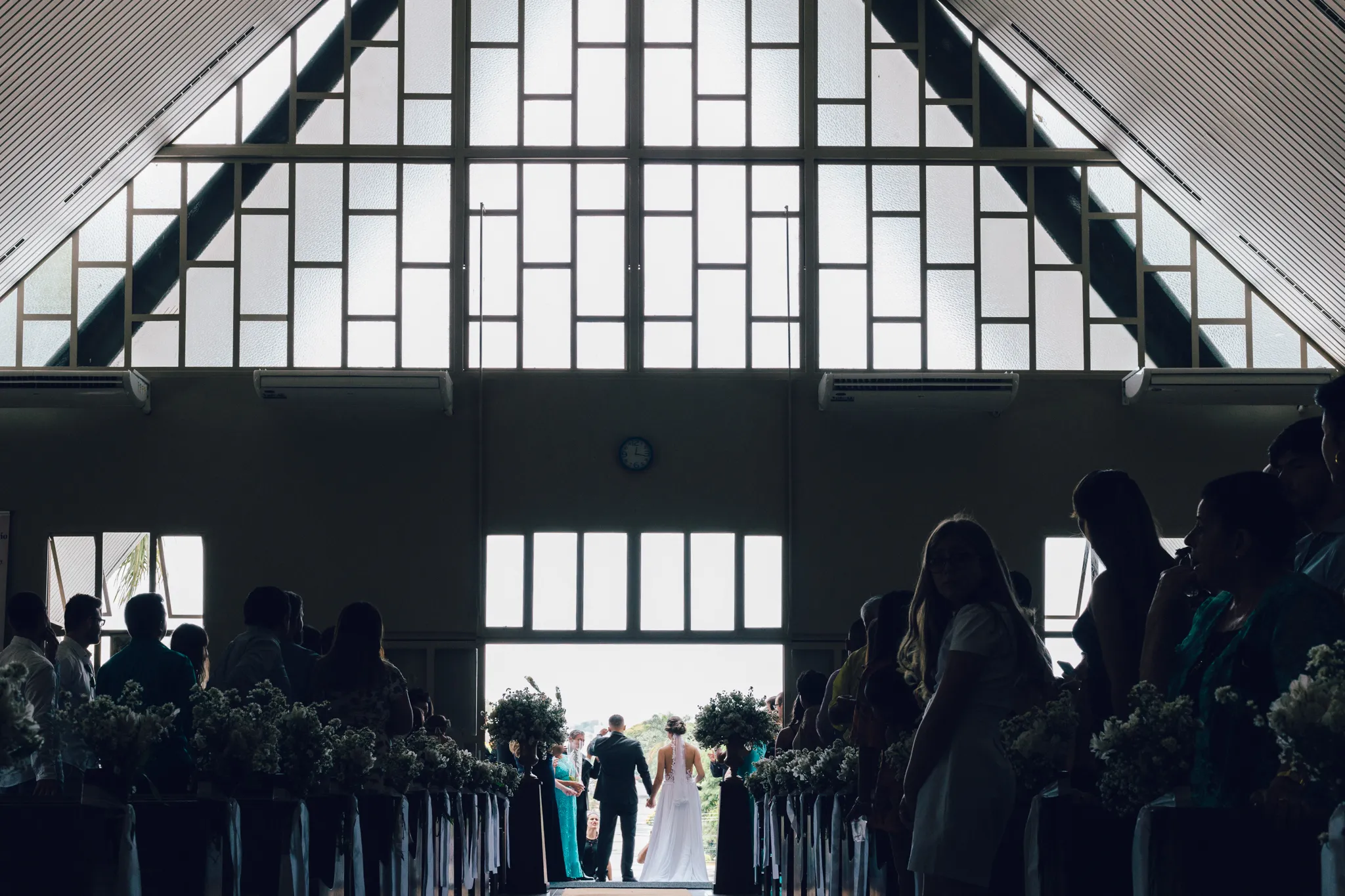 A igreja vista do fundo, em contraluz, com os noivos entrando pelo corredor entre os convidados