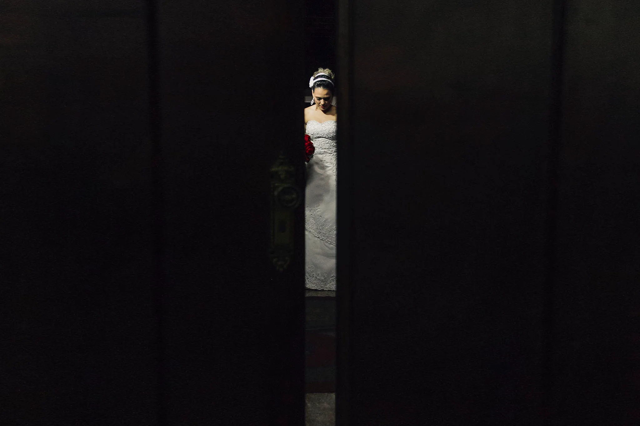 Pela fresta entre as duas folhas da porta, a noiva de vestido branco e buquê de rosas vermelhas, de olhos baixos, esperando a hora de entrar