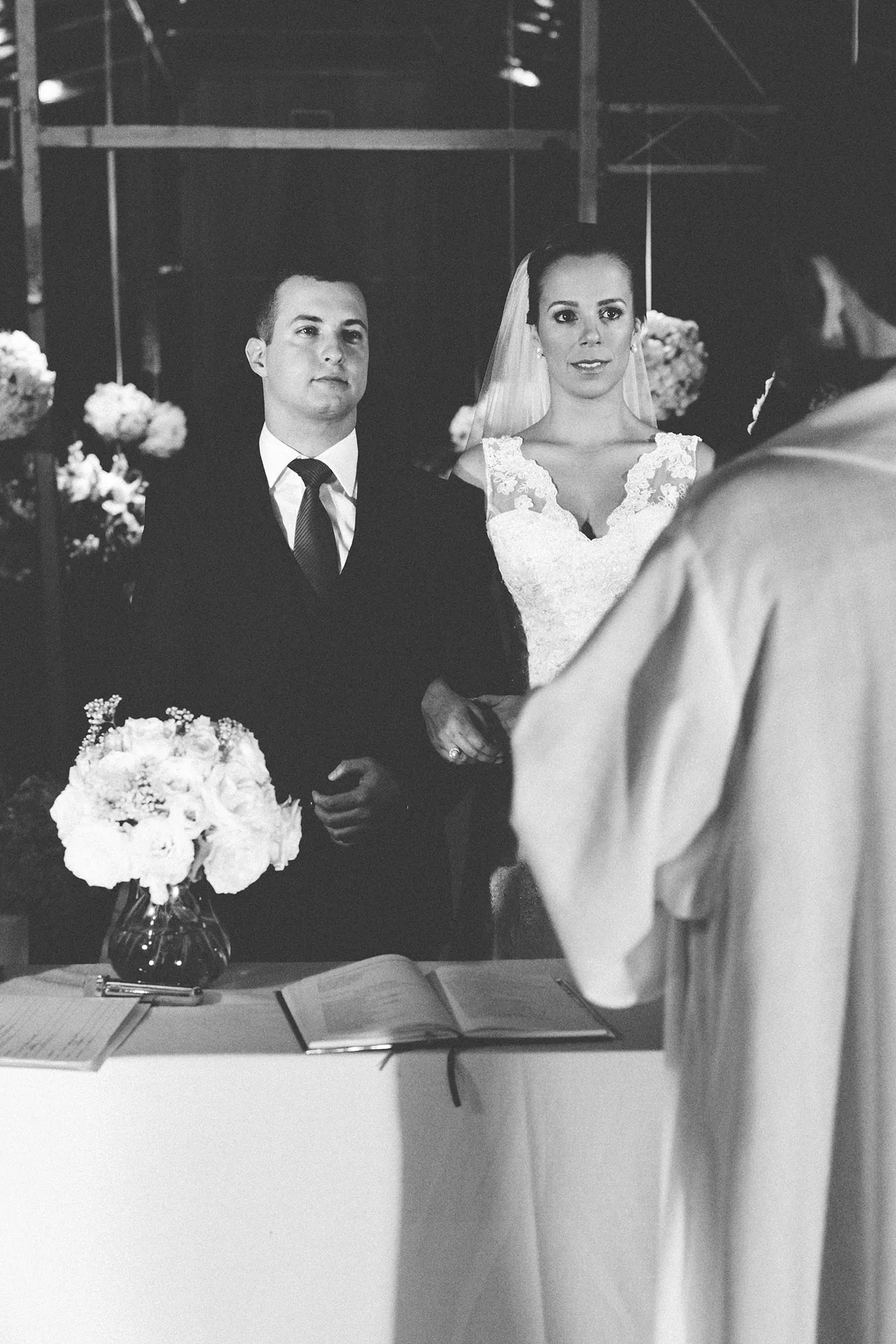 Os noivos lado a lado no altar, durante a cerimônia, ela sorrindo, em preto e branco